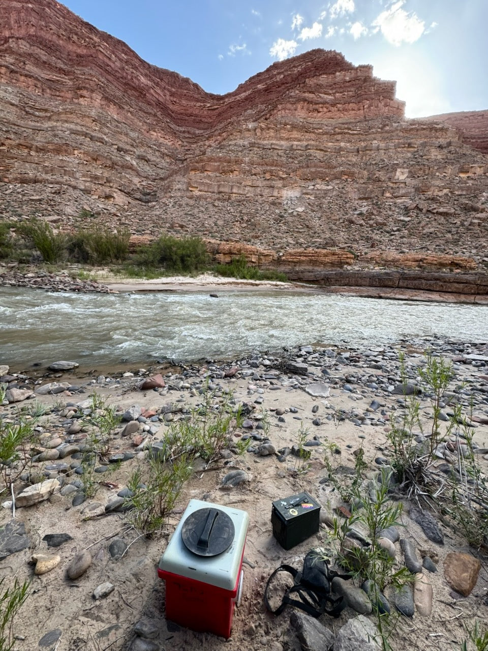 A 4CRS Rental Toilet stands on rocky ground beside a river, with layered canyon cliffs in the background under a partly cloudy sky.
