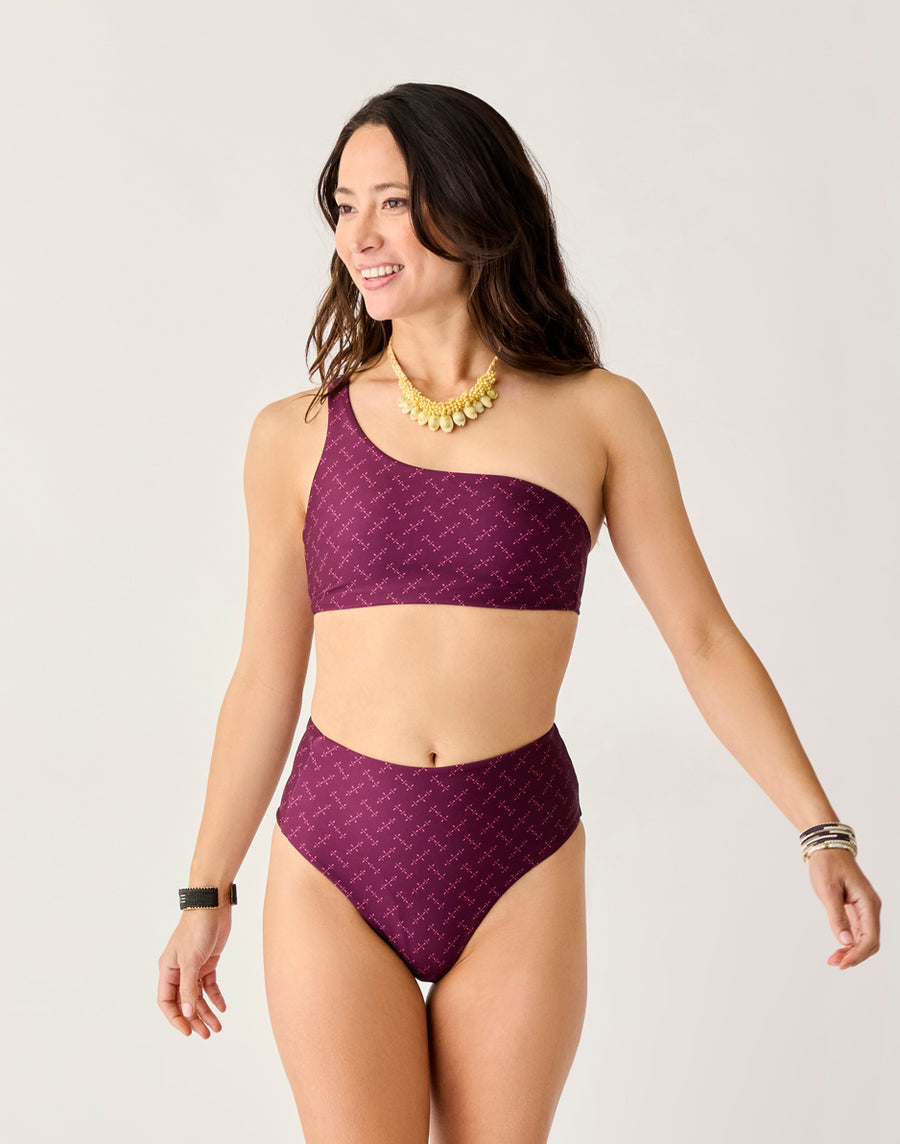 Woman wearing the Carve Lani Pocket Compression Bottom bikini, featuring a one-shoulder top and high-waisted fit in maroon with a subtle pattern. Accessories include gold jewelry and a smartwatch, standing against a plain light background.