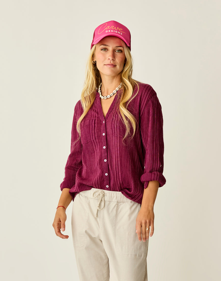 A woman stands against a plain background wearing the Carve Dylan Gauze Tunic in magenta, paired with light pants, a pink cap, and a shell necklace.