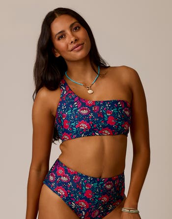 A woman models the Carve Bondi Compression Top, a blue and pink floral one-shoulder bikini crafted from recycled compression fabric, paired with a turquoise necklace—ideal for surf and swim adventures.