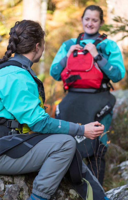 Two people in teal 4CRS Rental Drysuits and safety gear sit on rocks in a forested area, talking and smiling outdoors.