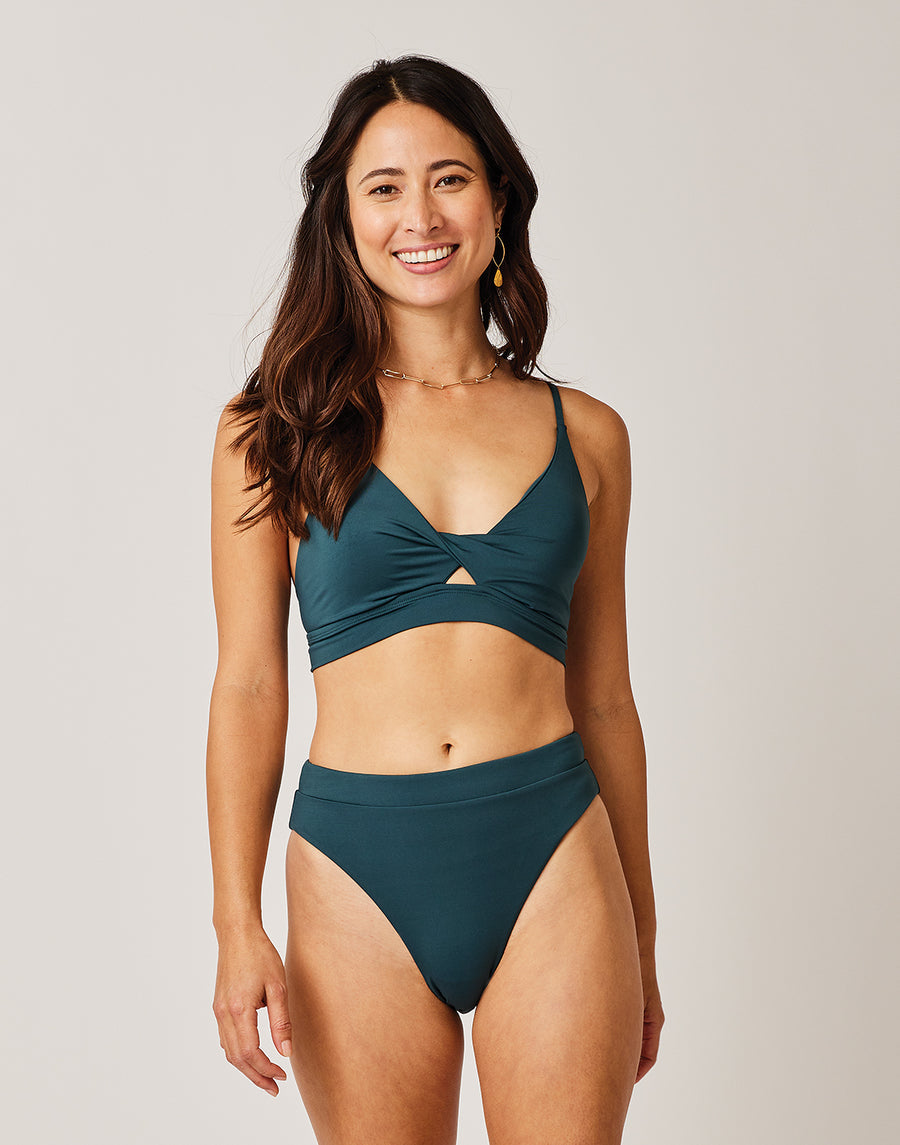 A woman stands smiling against a light background, wearing the Carve Elba Top by Carve—a dark teal, seamless bralette bikini top paired with matching high-waisted bottoms.