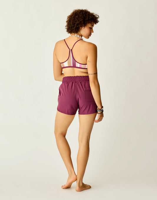 A woman stands barefoot facing away, wearing Carve's Bali Short and a striped sports bra, set against a plain light background.