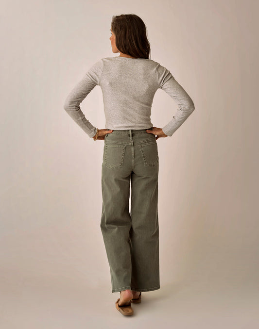 A woman stands facing away with her hands on her hips, wearing a light grey long-sleeve top and olive green Audrey Wide Leg Pant by Carve, paired with brown flats against a plain background.