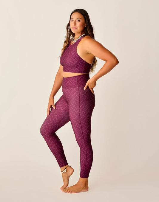 A woman poses barefoot in a side view, wearing Carve's Saluda Compression Tight and matching maroon athletic crop top, both made from recycled polyester with a subtle pattern, against a plain light background.