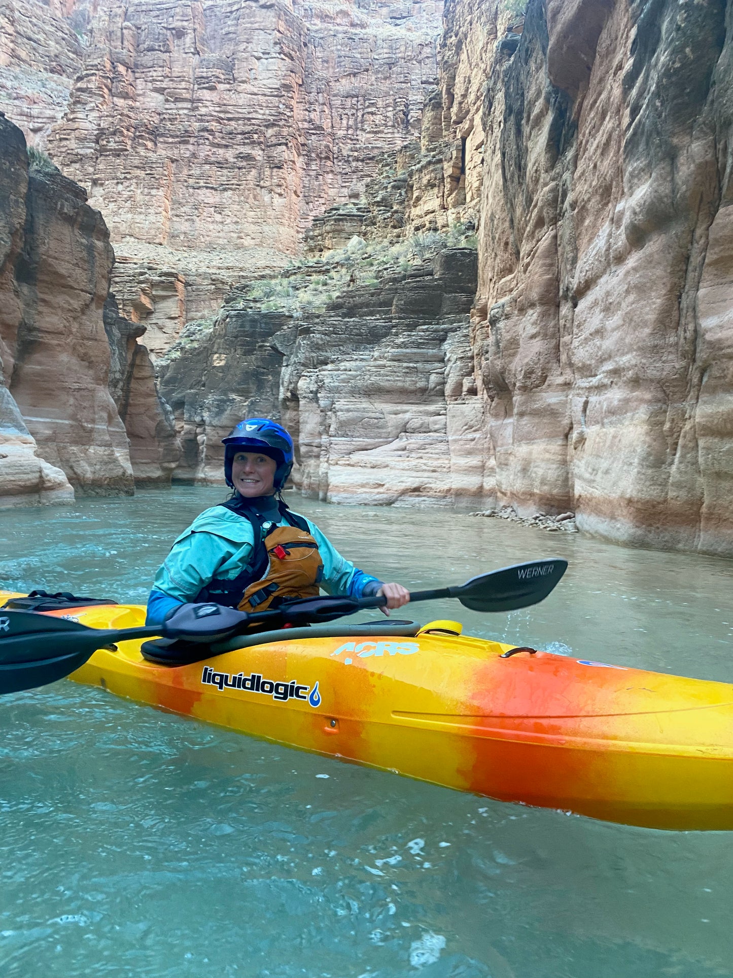 Outfitted in a helmet, life jacket, and 4CRS Grand Canyon Rental Drysuit, a paddler navigates a yellow kayak through a narrow, rocky canyon with turquoise water.
