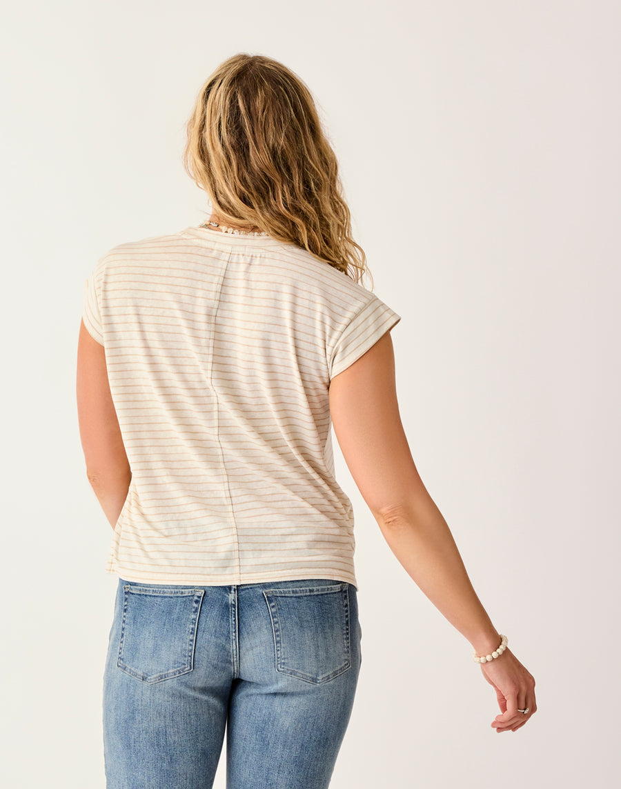 A person with wavy blonde hair is seen from the back, wearing Carve's Astrid Hemp Tee—a short-sleeve striped shirt—paired with blue jeans, set against a plain light background.