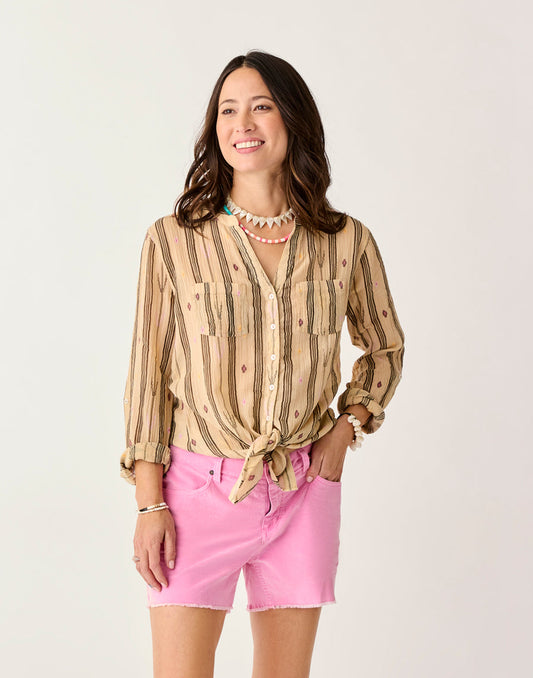 A woman wears the Carve Dylan Gauze Tunic, a tan and brown striped shirt tied at the waist, paired with pink shorts and layered necklaces, standing against a plain light background.
