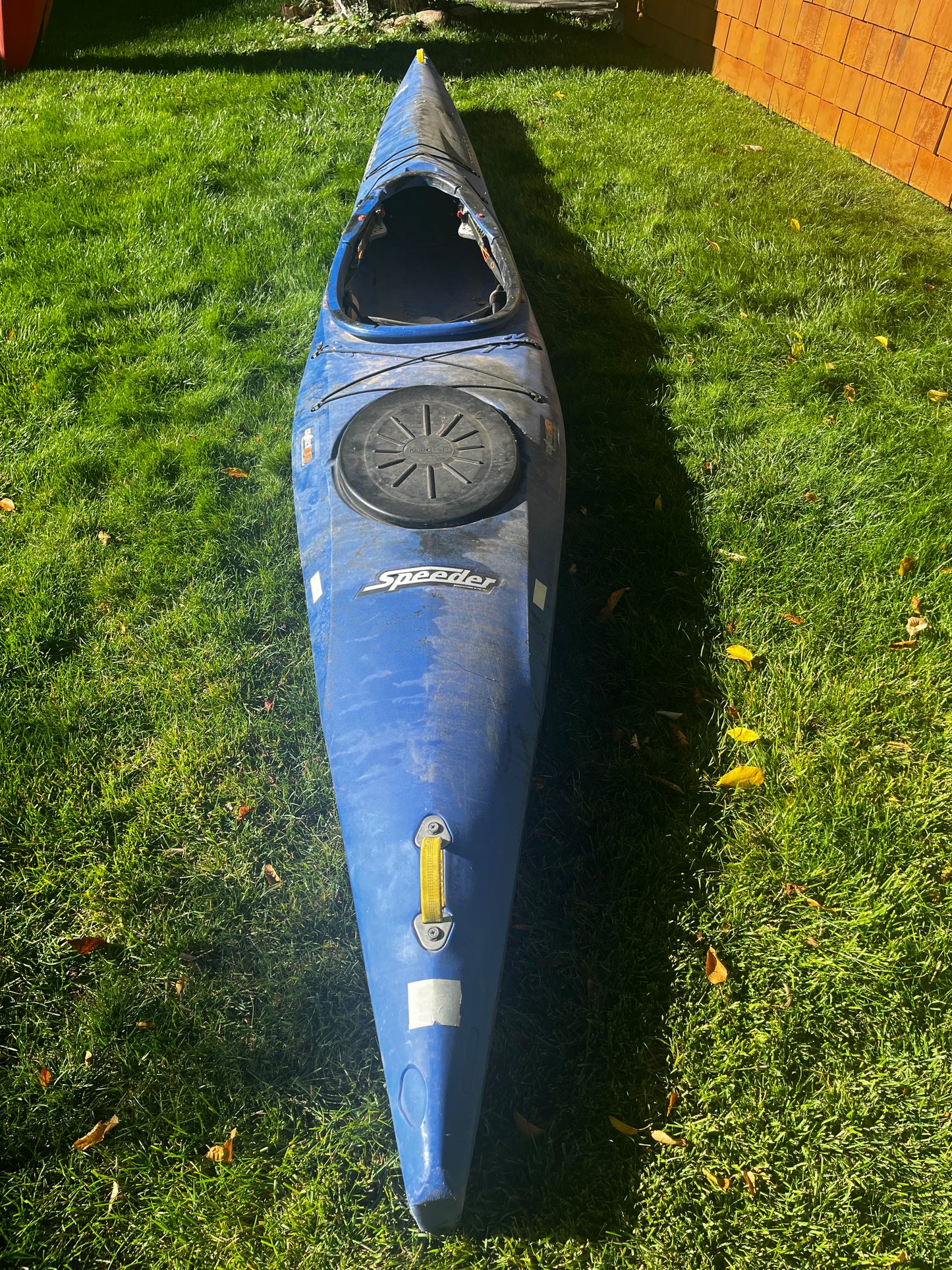 A blue Pyranha Speeder 14' kayak from 4Corners Riversports with a covered storage hatch rests on green grass next to a wooden building.