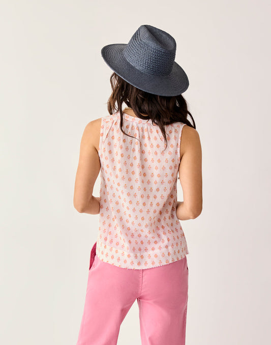 A woman faces away from the camera wearing the Carve Dylan Tank by Carve—a sleeveless white organic cotton top with a small pink pattern—paired with pink pants and a blue wide-brimmed hat, against a plain background.