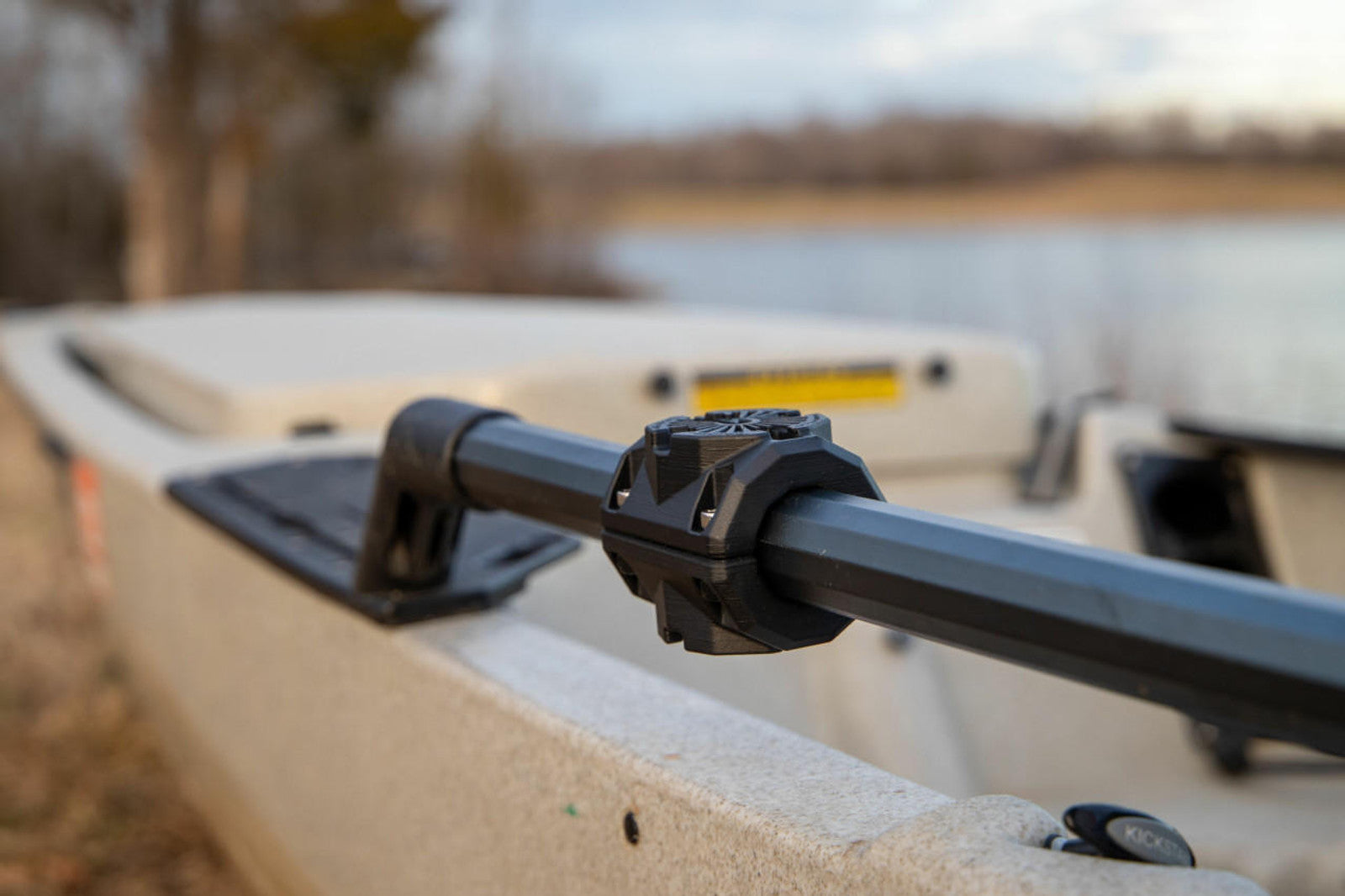 Close-up of a YakAttack MightyMount Duo Rail Adapter securing a paddle to the side of a beige kayak, with a blurred lake and trees in the background.