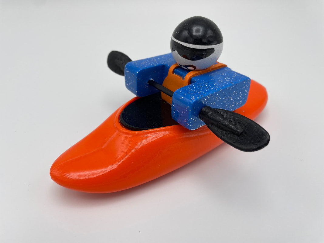 A toy figure dressed as a kayaker in blue gear and a black helmet skillfully navigates the Foamie Friends Foamie Mini Playboat, featuring an innovative hull design. Seated in an orange kayak, the figure confidently maneuvers the river with a black double-ended paddle, ready for any River Running adventure.