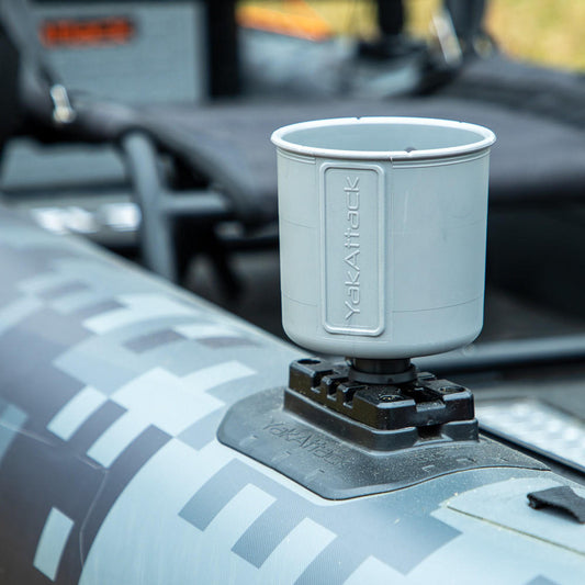 A YakAttack MultiMount Cup Holder in gray is attached to a camouflage kayak rail, with kayak seating visible in the background.