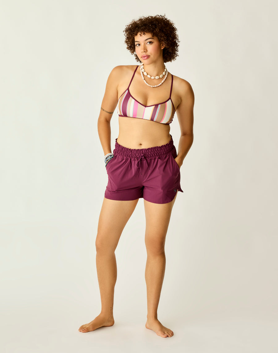 A woman stands barefoot against a plain light background wearing the Carve Bali Short in purple, paired with a striped bikini top, a beaded necklace, and bracelets.