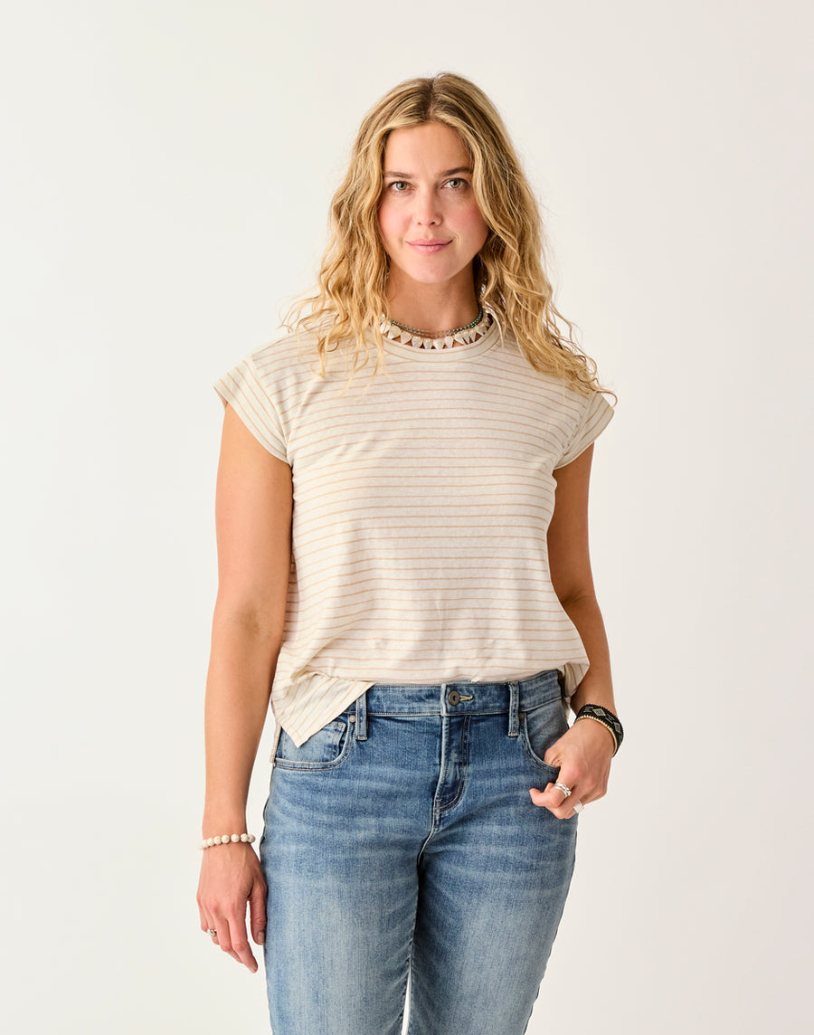 A woman with wavy blonde hair wears the Carve Astrid Hemp Tee, a short-sleeve, light-colored striped shirt, paired with blue jeans and stands against a plain light background.