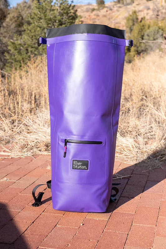 A purple, portable River Station Gear Dry Thwart Bag - V2 standing upright on a brick surface with a natural, grassy background.