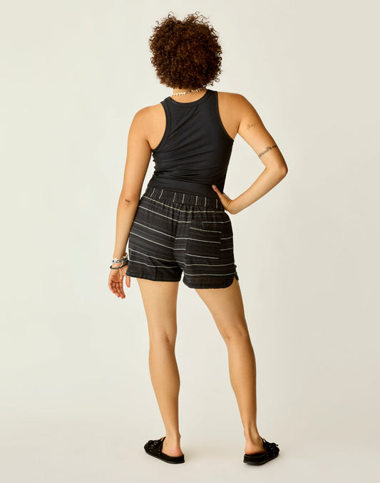 A person stands facing away, wearing Carve's black sleeveless top, Rhett Stitched Shorts in black and white stripes, and black slide sandals against a plain background.