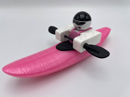 A Foamie Mini Creeker from the Foamie Friends brand features a small toy figure wearing a helmet in a pink kayak, holding black paddles against a plain background. This figure is ideal for imaginative river play adventures.