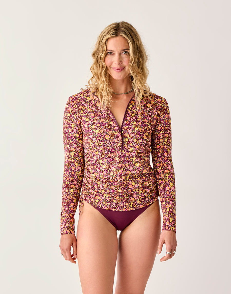 A woman faces forward against a plain background, wearing the Carve Cruz Rashguard with long sleeves and a floral print, paired with maroon bikini bottoms.
