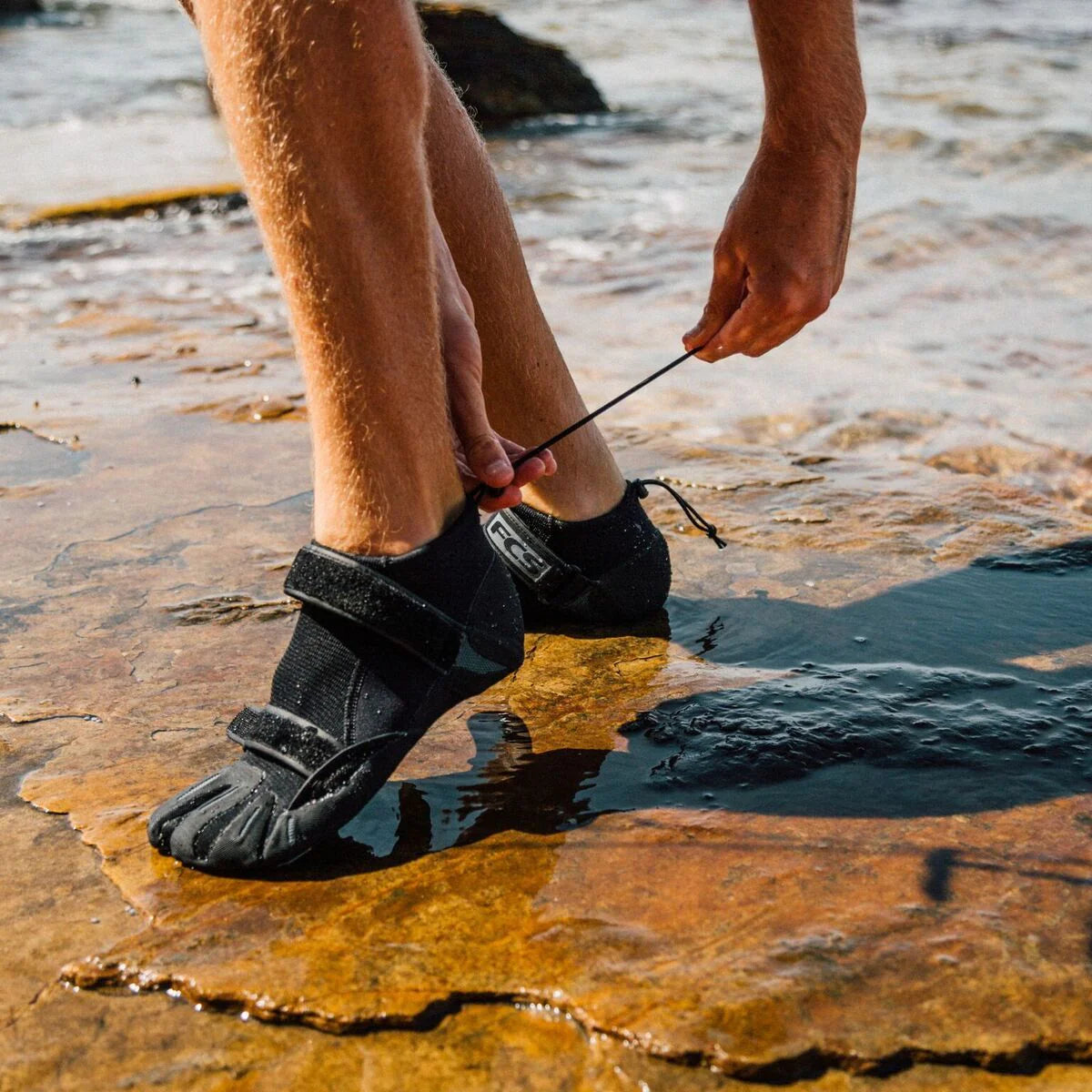Wearing FCS SP2 Reef Booties with ergonomic Velcro straps, someone secures the fit while standing on wet, rocky terrain near shallow water.
