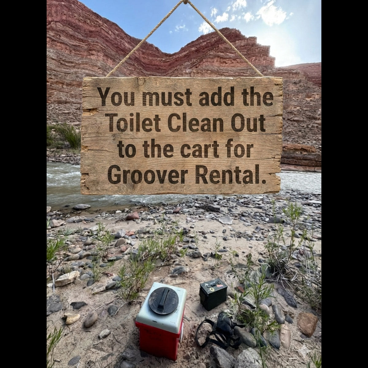 A 4CRS portable toilet and gear rest on rocky ground by a river and red cliffs. A wooden sign says, "You must add the 4CRS Toilet Clean Out to the cart for Groover Rental.