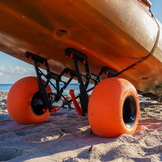 Close-up of a kayak with a YakAttack TowNStow Bunkster Kayak Cart w/ Sand Wheels, featuring large orange wheels and adjustable uprights, resting on the sandy beach near the ocean.