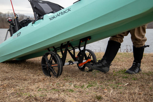 A person in waders pulls a green kayak on a YakAttack TowNStow Bunkster Kayak Cart with sand wheels near a lake.