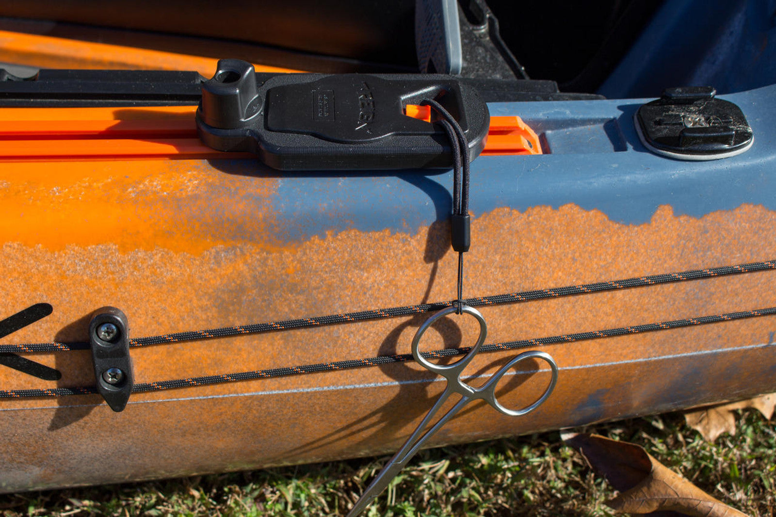 Close-up of an orange and blue kayak featuring the YakAttack Track Mount Retractor with metal forceps attached to the track—perfect for organizing kayak fishing gear. Grass and leaves cover the ground below.