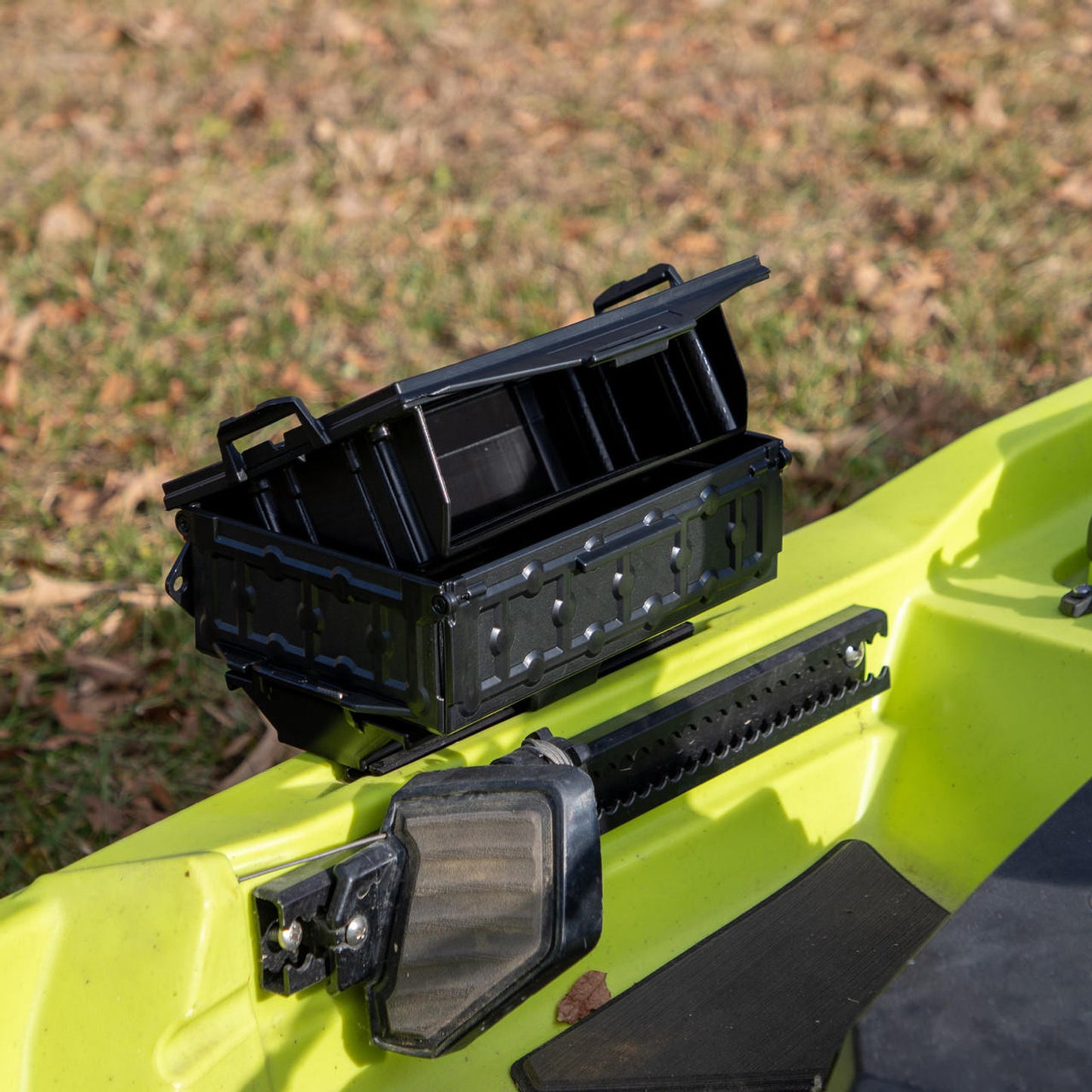 A YakAttack TracPak with PicPocket QuickDraw is attached to a yellow kayak, its lid open. Grass and dried leaves are visible in the background.