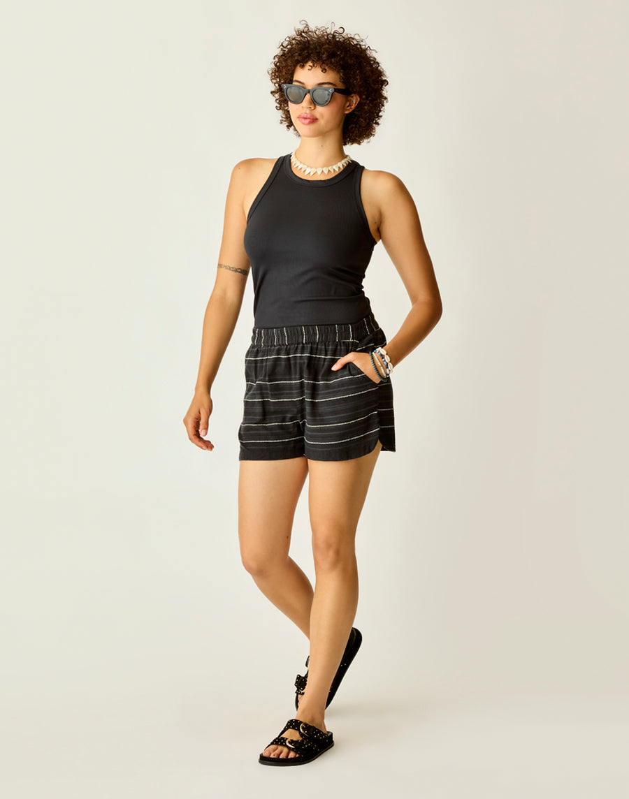 A person with curly hair wears sunglasses, a black sleeveless top, Carve's Rhett Stitched Shorts, black sandals, and accessories, standing against a plain light background.