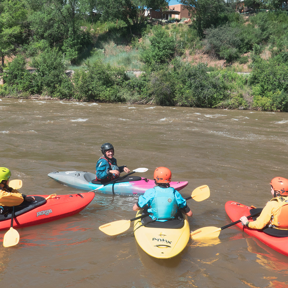 Four kayakers in different colored kayaks are gathered on a river. The scene is surrounded by lush greenery and a two-story house is visible in the background.