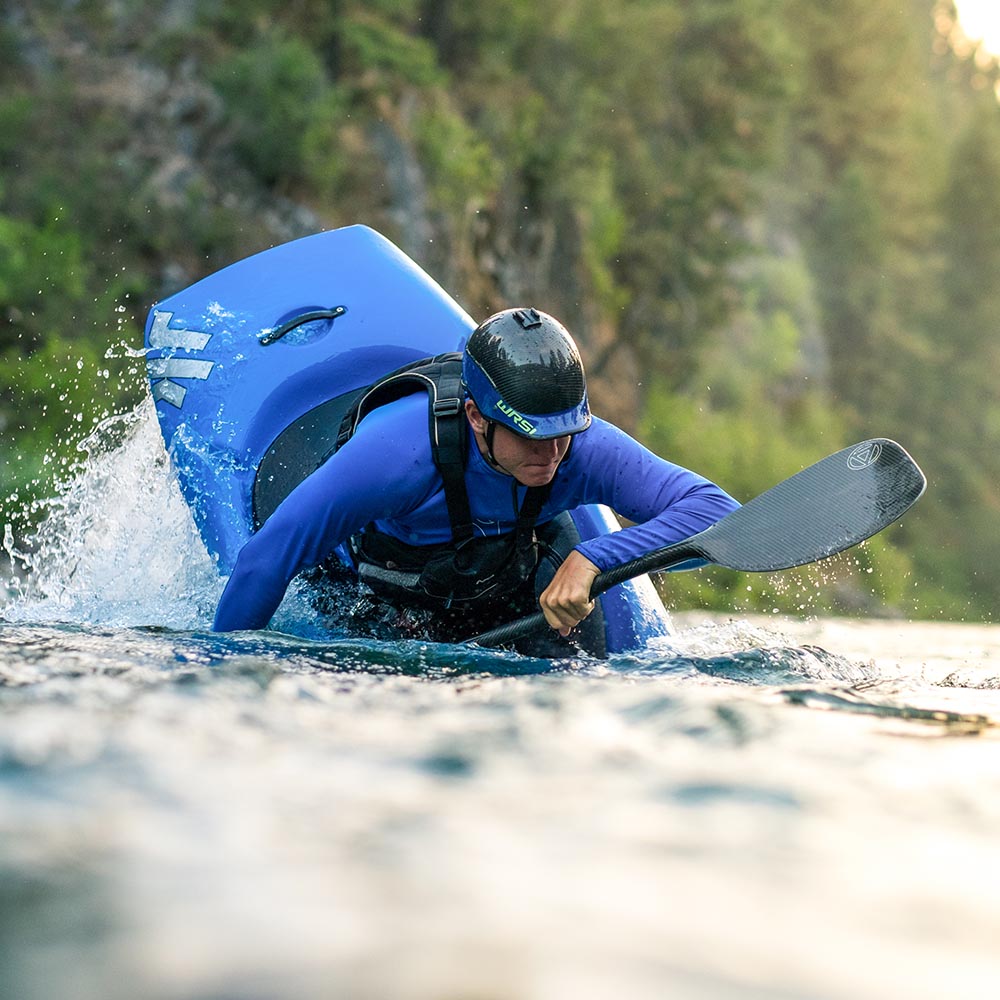 Person in a helmet and life jacket performing a kayaking maneuver on a river with splashing water and trees in the background.
