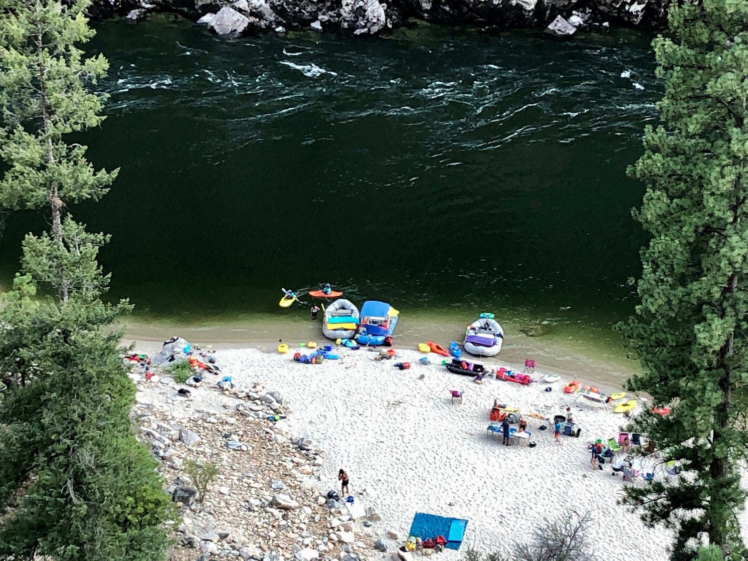 Aerial view of a sandy riverbank with inflatable rafts, people, and camping gear along the water’s edge, surrounded by trees and rocky shoreline.