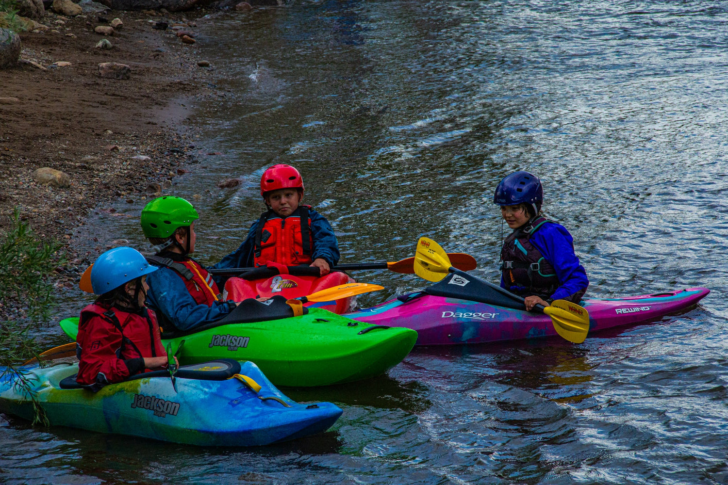 Four people in helmets and life jackets sit in colorful kayaks on a river near the shore, appearing to converse or prepare for an activity.