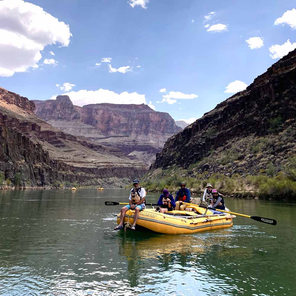 A group of people sit on a yellow inflatable raft, paddling on a calm river surrounded by rocky cliffs and a clear sky overhead.