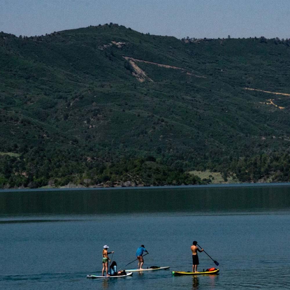 Three individuals paddleboarding on a calm lake with green, tree-covered hills in the background under a clear sky.