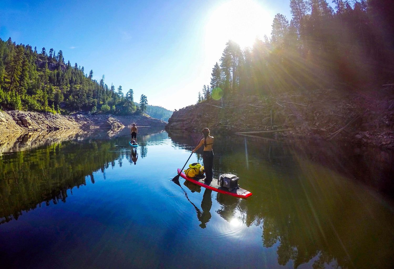 Two people paddleboard on a calm, reflective lake surrounded by trees and rocky banks under a clear, sunny sky.