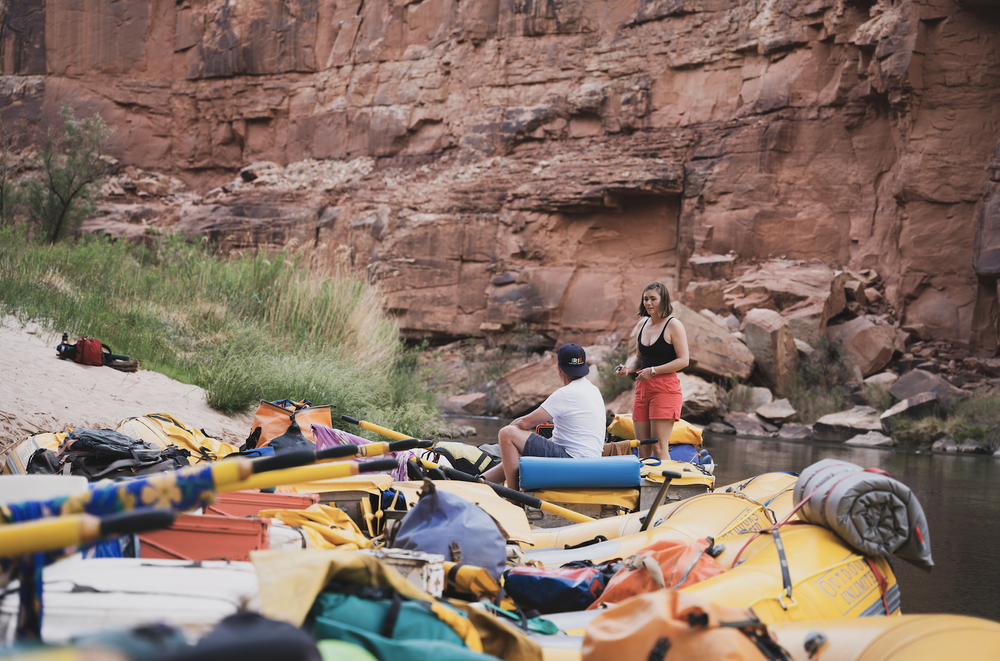 Two people stand near rafts loaded with gear on a riverbank, with a rocky, red canyon wall in the background.