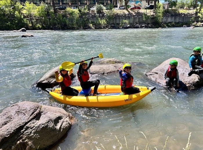 Three people in a yellow inflatable kayak paddle on a river while two others sit on rocks nearby, all wearing helmets and life jackets.
