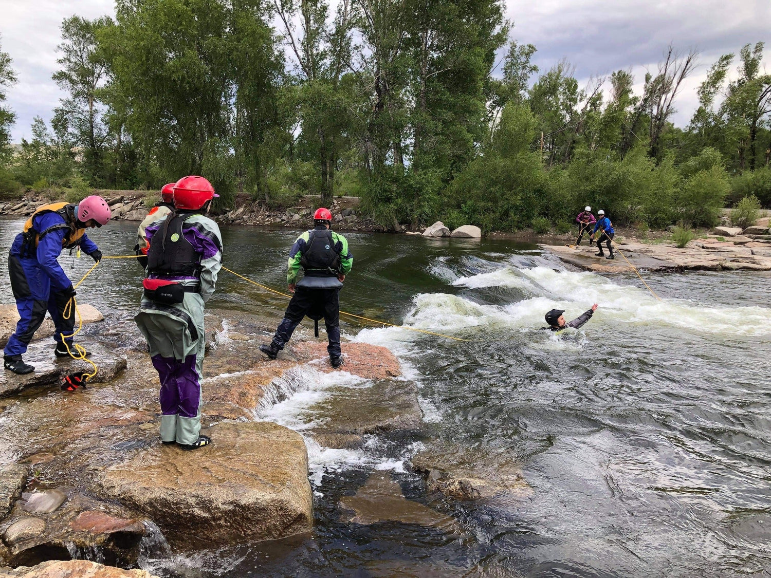 A group of people in safety gear perform a river rescue drill, with one individual in the water holding a rope, while others stand on the rocks guiding the practice.