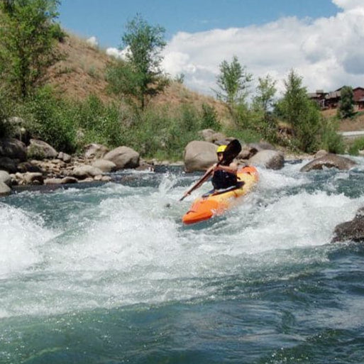 A person wearing a helmet kayaks through whitewater rapids on a river with rocky banks and trees under a partly cloudy sky.