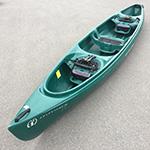 A green three-seat kayak is placed on a concrete surface with two black seats fitted with storage compartments.