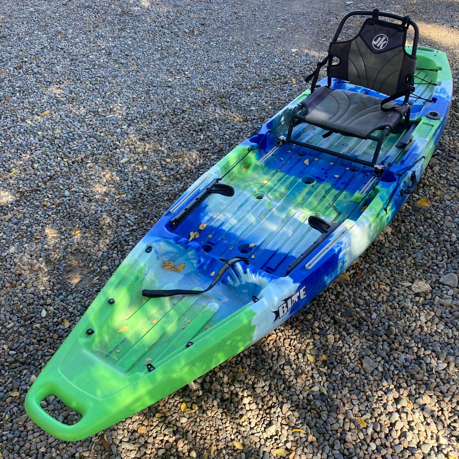 A green, blue, and white sit-on-top kayak is positioned on a gravel surface, featuring a padded seat with a backrest and multiple storage compartments.
