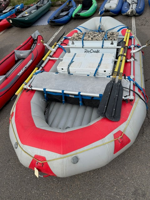 A red and gray inflatable raft with oars, a cooler labeled "RioCraft," and gear strapped inside, resting on a paved surface among other rafts.