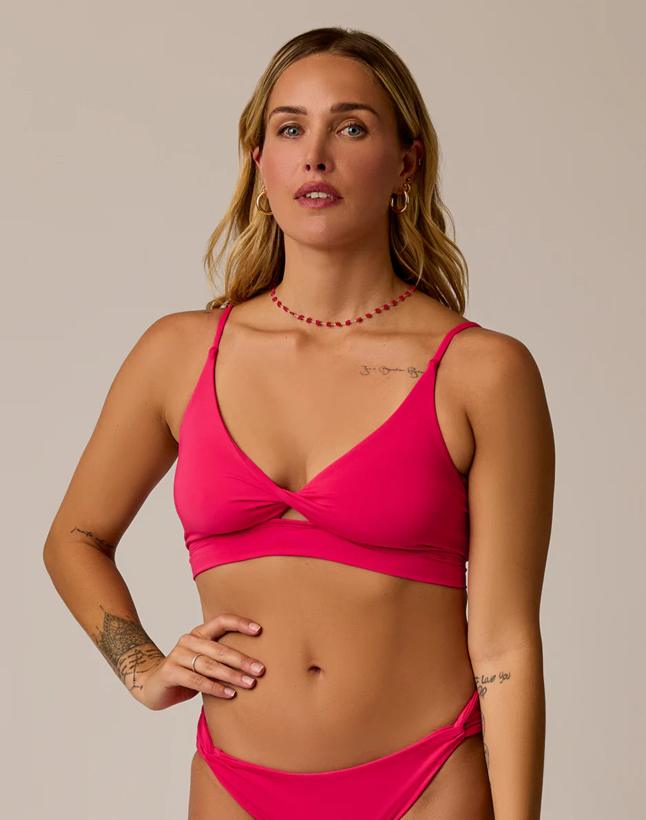 A woman wearing the Carve Elba Top by Carve, a bright pink twisted front bikini with adjustable straps, stands against a plain background with one hand on her hip, showing arm tattoos and minimal jewelry.