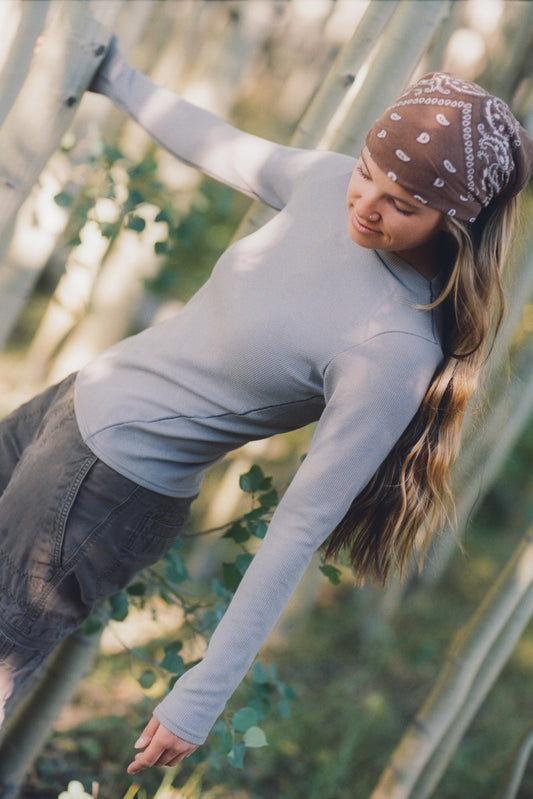 A person with long hair and a bandana leans sideways among trees, wearing the Allover Mesa Long Sleeve W's shirt with adjustable-strap overalls and pockets, looking down neutrally.