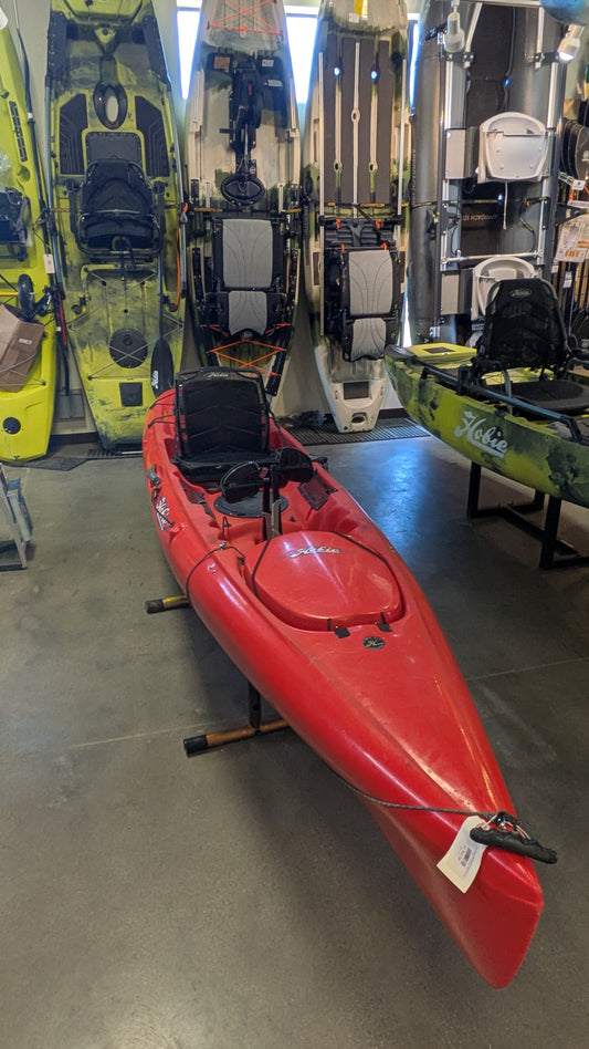 A Consignment Hobie Mirage Revolution 13 kayak from 4Corners Riversports is indoors on a stand, surrounded by upright kayaks and paddle sport gear.