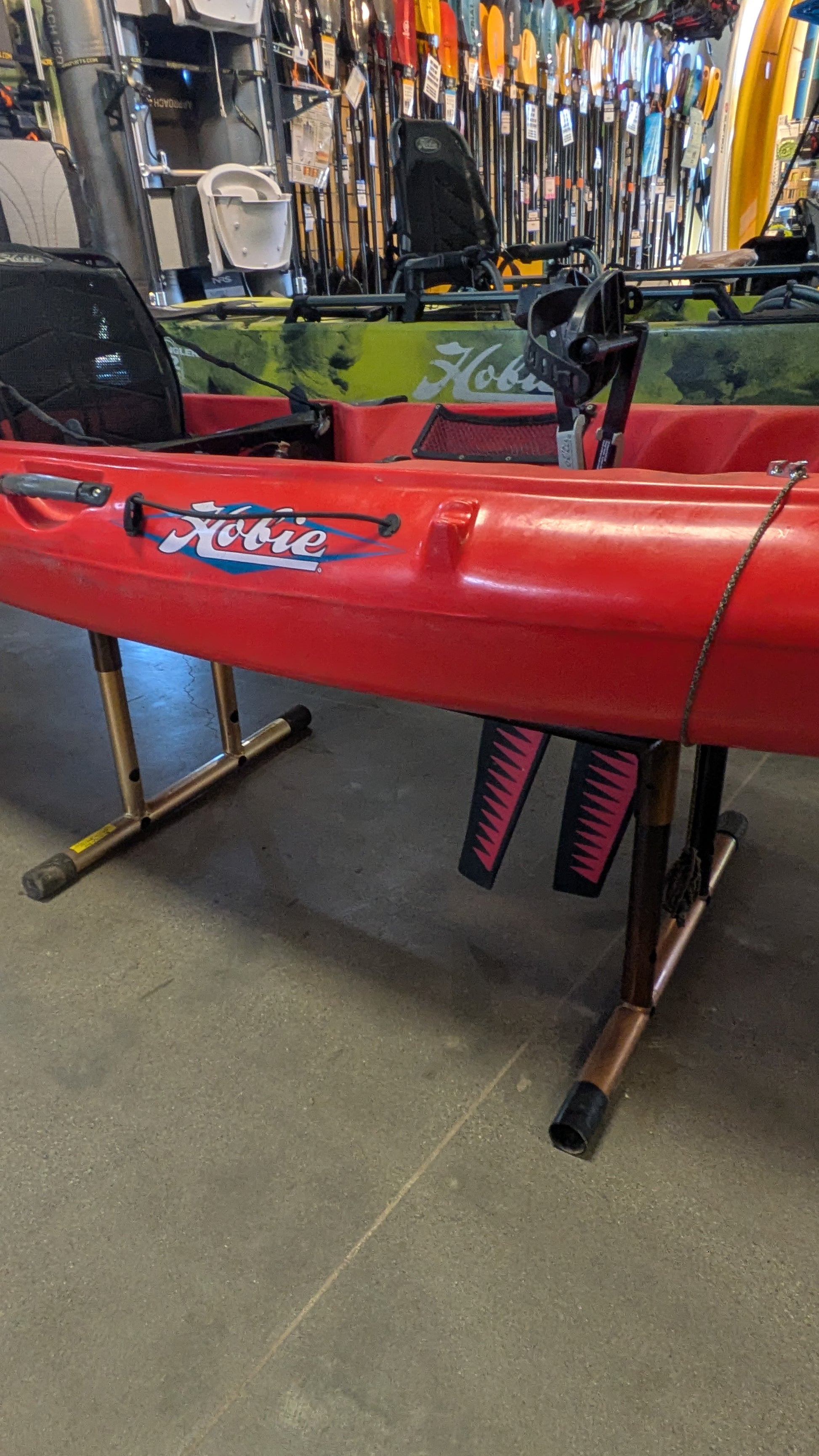 A Consignment Hobie Mirage Revolution 13 kayak from 4Corners Riversports, featuring kick-up fin technology, is displayed on a stand in a store with paddles and other kayaks visible in the background.