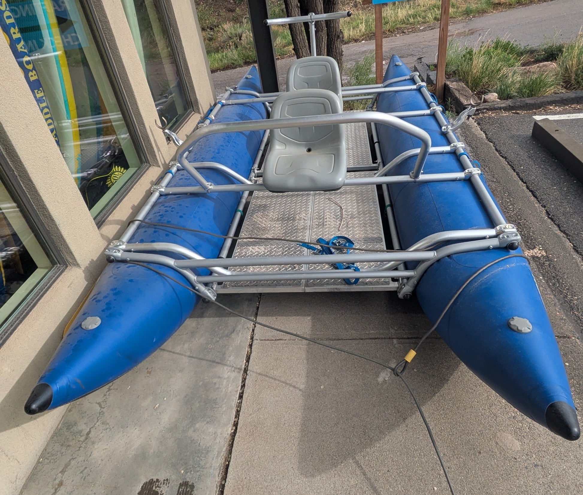A 16' Cataraft Saturn from 4Corners Riversports with a metal frame and two seats is parked on a sidewalk near a building beside a "Rafting Check In" sign.