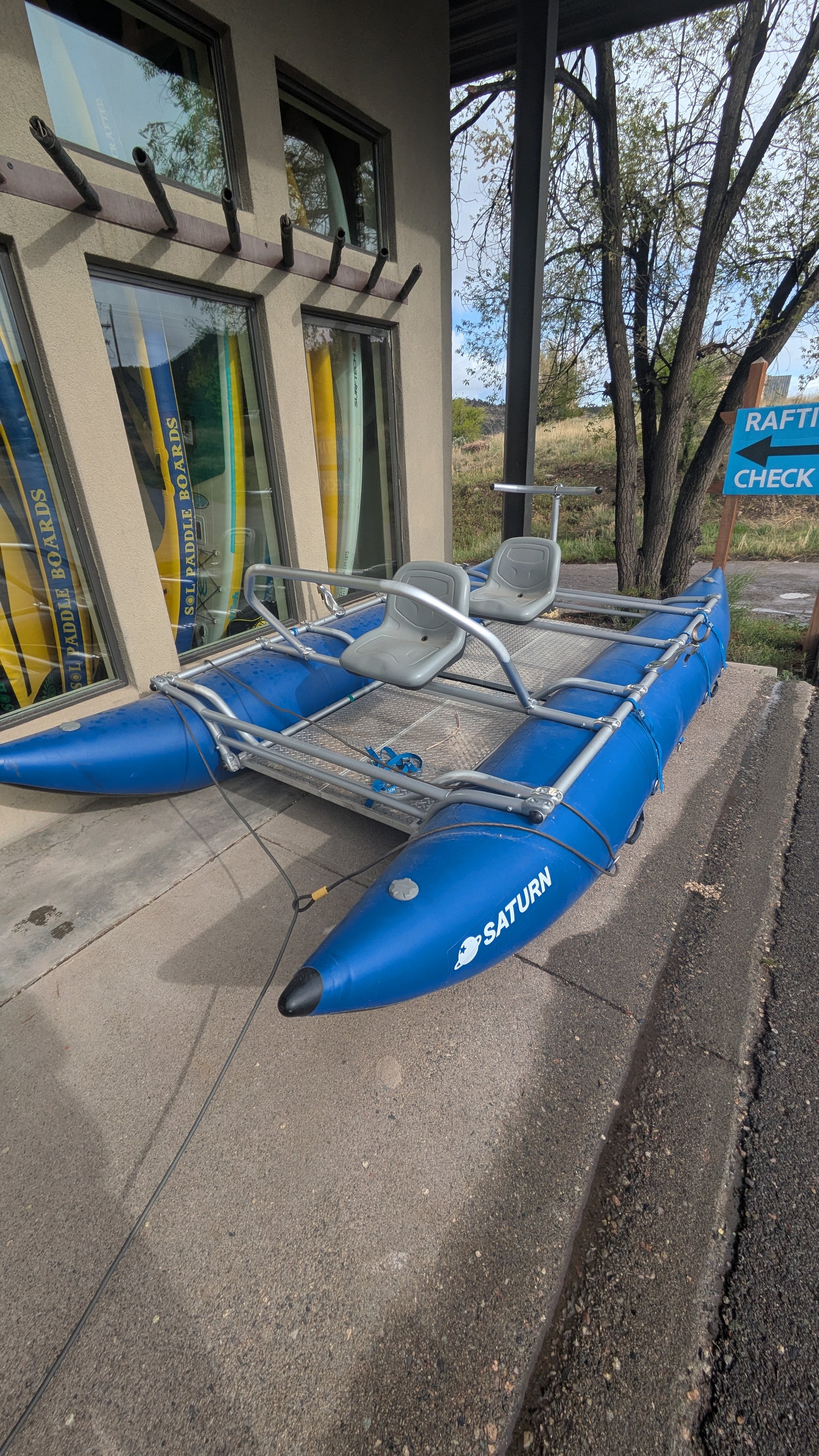 A 16' Cataraft Saturn by 4Corners Riversports with two seats is parked outside a building with large windows and a "Raft Rentals" sign nearby.